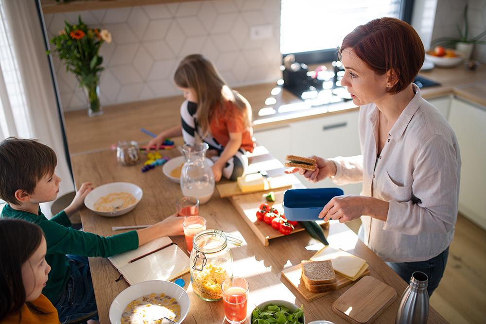 A woman prepares sandwiches in a bright kitchen while three children sit at the table, eating and playing. The table has food, drinks, and toys. Sunlight streams through the window.