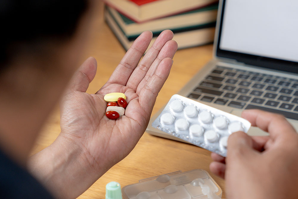 A person holding several pills in one hand and a blister pack in the other, sitting at a desk near a laptop, books, and medication containers.