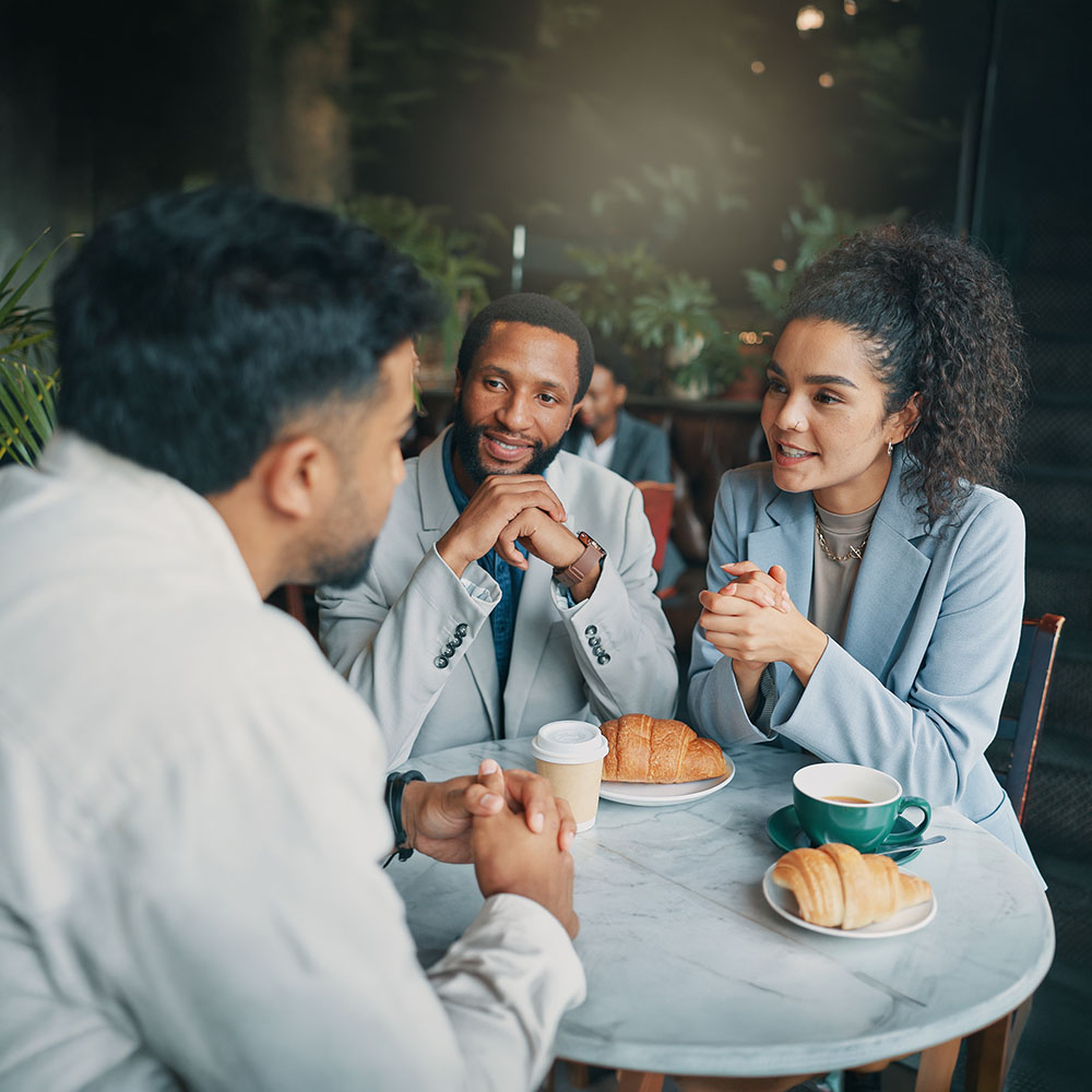 Three people sit at a round café table, talking and smiling. One of them, who has hearing loss, leans in to better catch the conversation as they enjoy their time together. Two have croissants and coffee in front of them. The setting is cozy with green plants in the background.