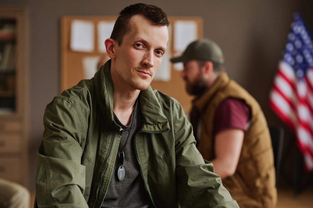 Man in a green jacket with dog tags sits indoors, looking at the camera, while another man in a cap and brown vest sits in the background near an American flag.