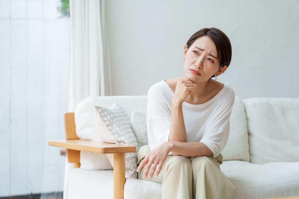 A woman in a white shirt sits on a sofa, resting her chin on her hand and looking thoughtfully into the distance.