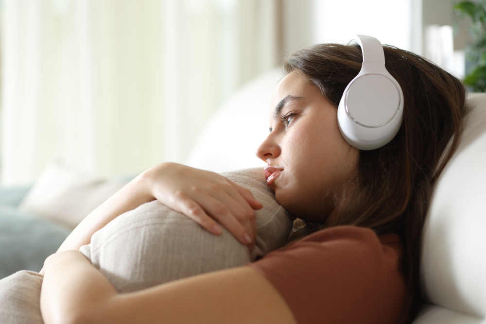 A young woman wearing headphones hugs a pillow while sitting on a couch, looking thoughtful and sad.