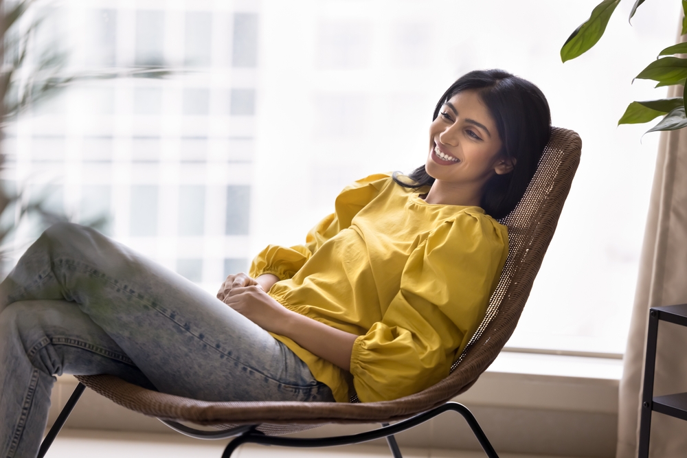 A woman in a yellow blouse and jeans smiles while relaxing in a chair near a window, with greenery and city buildings in the background.