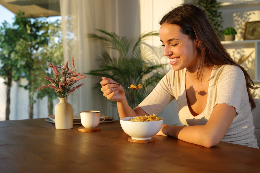 A woman sits at a wooden table, smiling as she eats cereal from a bowl. There is a mug and a vase with flowers on the table, with plants and sunlight in the background.