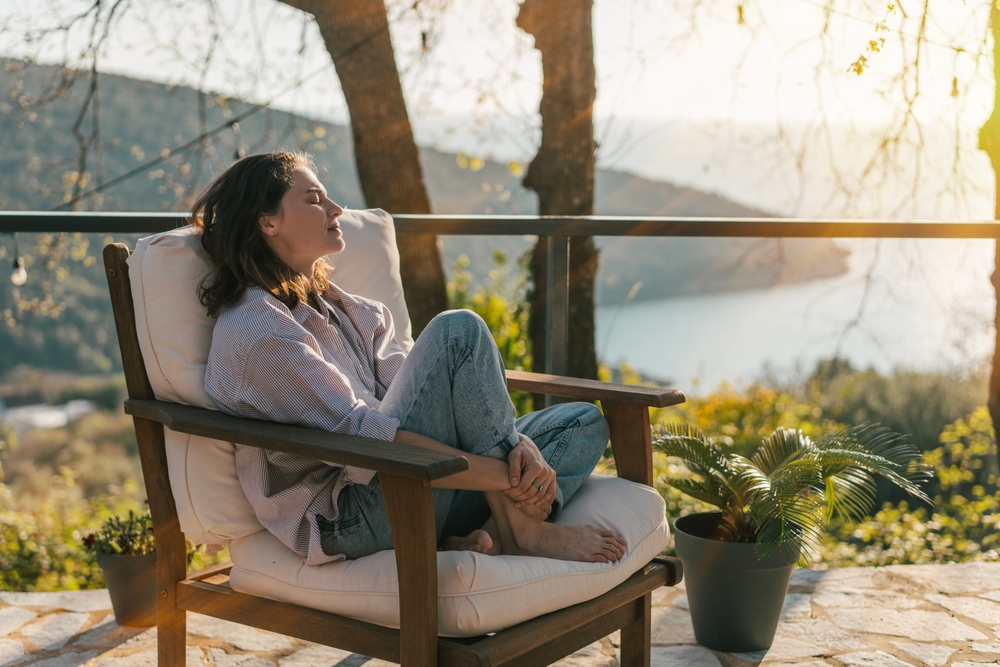 A woman relaxes in a cushioned chair on a patio, eyes closed, with sunlight on her face. Trees, hills, and a body of water can be seen in the background, along with a potted plant nearby.