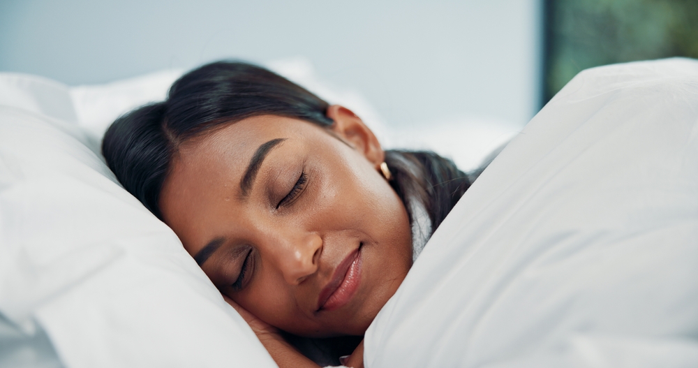 A woman with dark hair lies on her side in bed, eyes closed and smiling softly, covered by a white blanket.