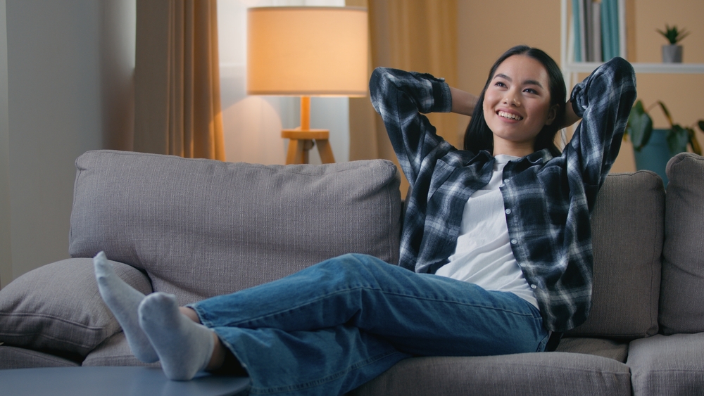 A woman sits on a couch with her legs crossed and hands behind her head, smiling and relaxing in a cozy living room with warm lighting and a lamp in the background.