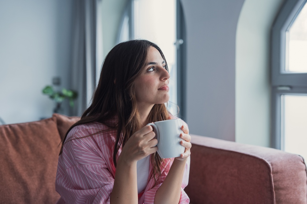 A woman in a pink robe sits on a couch, holding a white mug with both hands, and gazes thoughtfully out of a window.