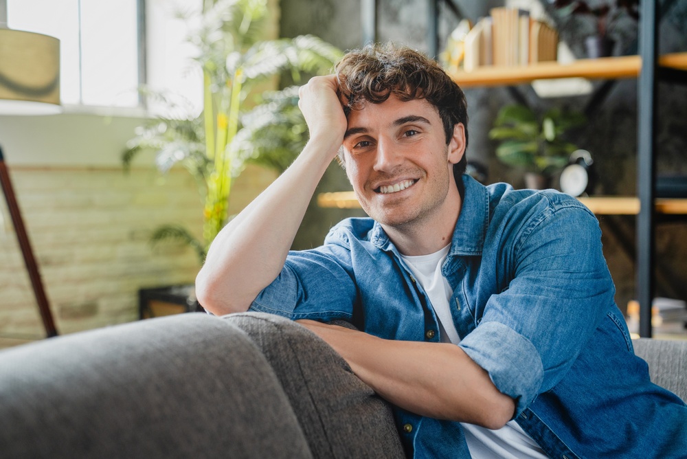 A young man with curly hair and a denim shirt smiles while sitting on a gray sofa in a cozy, sunlit living room with plants and shelves in the background.
